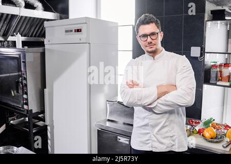 Porträt eines selbstbewussten jungen Küchenchefs in Brillen, der in Uniform mit gekreuzten Armen steht und die Kamera anschaut Stockfoto