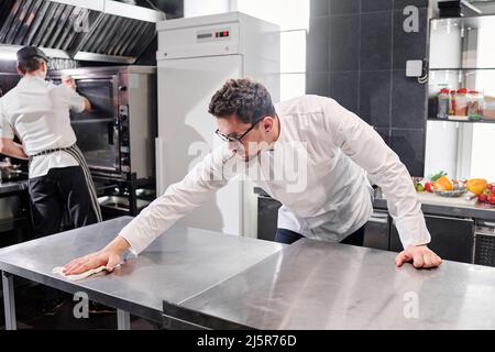 Koch in Uniform wischen Küchentisch nach dem Kochen mit seinem Kollegen Reinigung des Ofens nach der Arbeit in der Küche Stockfoto