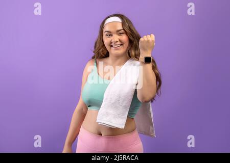 Frohes junges europäisches Plus size Weibchen in Sportbekleidung mit Handtuch zeigt nach dem Training einen Fittracker Stockfoto