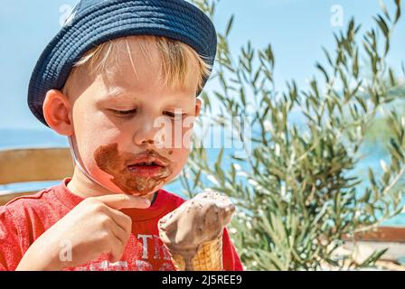 Blonde junge in panama Hut genießt essen Schokolade Eis immer schmutzig. Niedliches Kleinkind sitzt auf der Bank gegen das Meer im Urlaub in Omis Nahaufnahme Stockfoto