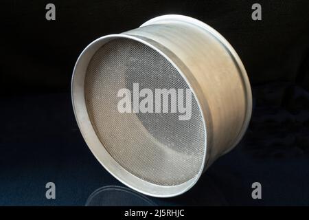 Ein Metallmehlsieb auf dem Küchentisch. Küchenutensilien auf schwarzem Hintergrund Stockfoto