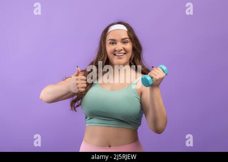 Die glad Millennial european Plus size Hündin in Sportswear trainiert mit Kurzhanteln und zeigt den Daumen nach oben Stockfoto