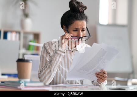 Weibliche Büromanagerin, die eine Brille trägt und Papierarbeiten macht, Augenprobleme hat und am Arbeitsplatz sitzt Stockfoto