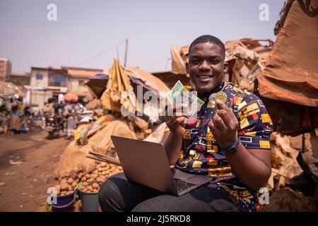 Afrikanischer Junge auf dem Markt mit FCFA-Francs und einer Bitcoin-Münze, Implementierung von Kryptowährungen in afrika Stockfoto