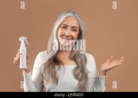 Lächelnde ältere Frau mit Sprühflasche für die Hautpflege oder Haarreparaturbehandlung auf braunem Studiohintergrund Stockfoto