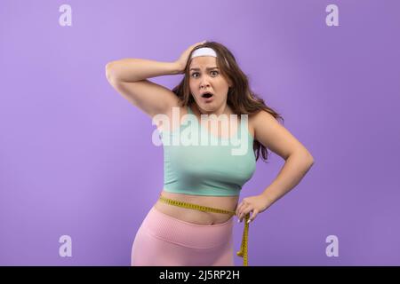 Überrascht traurig junge europäische plus size Frau in Sportbekleidung mit offenem Mund misst Taille mit Maßband Stockfoto