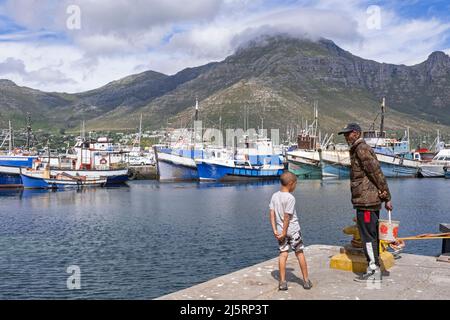 Älterer schwarzer Mann und Junge beobachten Fischerboote im Hafen von Hout Bay / Houtbaai in der Nähe von Kapstadt / Kaapstad, Westkap, Südafrika Stockfoto