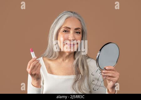 Ziemlich reife Dame, die im Spiegel schaut und Lippenstift auf braunem Studiohintergrund anwendet. Beauty- und Make-up-Konzept Stockfoto