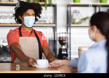 Lächelnder junger afroamerikanischer lockiger männlicher Kellner in Schutzmaske und Schürze, der dem Gast frischen Kaffee gab Stockfoto