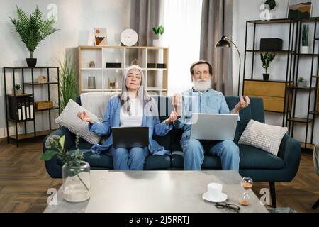 Die schöne reife Frau und ihr bärtiger Mann sitzen auf der Couch mit Laptops auf den Knien und meditieren mit geschlossenen Augen. Konzept der Entspannung und Harmonie. Stockfoto