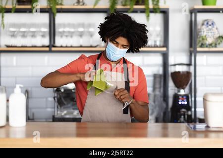 Beschäftigter, zweijähriger, schwarzer, lockiger Kellner in Schutzmaske und Schürze wischt hinter der Bar Glas mit Lappen ab Stockfoto
