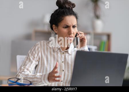 Verwirrte junge Geschäftsfrau, die mit einem Laptop über das Mobiltelefon spricht, Probleme mit dem Computer hat und im Büro arbeitet Stockfoto