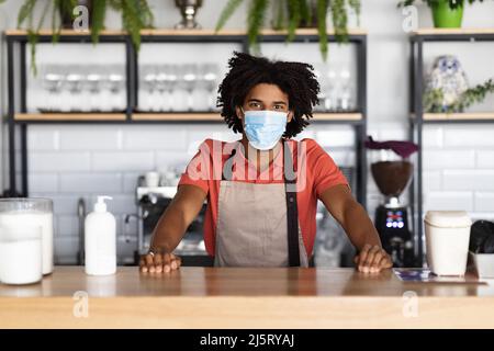 Glücklicher, selbstbewusster junger afroamerikanischer Lockkellner in Schutzmaske, Schürze steht an der Theke im Café Stockfoto
