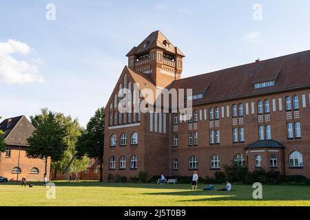 Hansa-Gymnasium in der Stadt. Bau der Sekundarschule in einer alten Architektur. Das Haus ist aus roten Ziegeln gebaut. Ein Park mit Menschen. Stockfoto