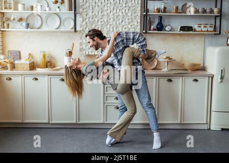 Glückliche tausendjährige europäische Frau und Mann tanzen zusammen in der modernen Küche Interieur, Profil Stockfoto