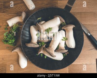 König Austernpilze mit frischen Kräutern in schwarzer Eisenpfanne auf einem hölzernen Schneidebrett, das mit wenig verziert ist Stockfoto