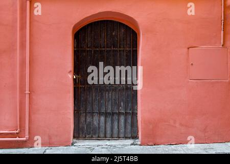 Schwarze Metallbogentür mit Kette und Vorhängeschloss an der hellrosa Wand eines alten Hauses in der Altstadt von Sanremo, Imperia, Ligurien, Italien geschlossen Stockfoto