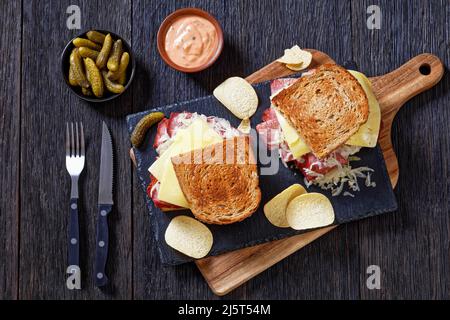 reuben Sandwich mit Roggenbrot dünn geschnittenes Corned Beef, Sauerkraut, russisches Dressing und geschmolzener Käse an Bord mit Gurken und Kartoffelchips, auf d Stockfoto