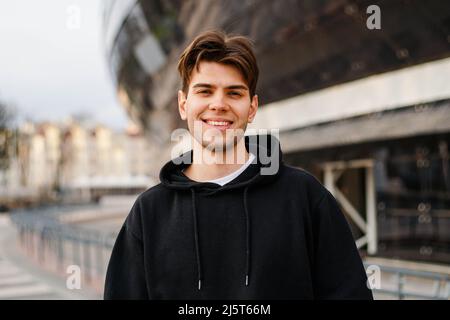Stadtportrait eines lächelnden Hipster-Mannes mit schwarzem Hoodie. Stockfoto