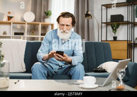 Porträt eines glücklichen Mannes mit grauem Bart reifer Mann in Freizeitkleidung, hält das Smartphone in den Händen, sitzt auf der Couch, entspannt im Wohnzimmer zu Hause, und Blick auf das Telefon in der Freizeit. Stockfoto