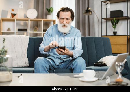 Videoanruf Ferngespräch Social Networks Konzept. Ein älterer Mann mit einem überraschenden Gesichtsausdruck sitzt zu Hause auf dem Sofa und hat Videoanruf mit seinem Smartphone, um mit Freunden zu reden und Spaß zu haben Stockfoto