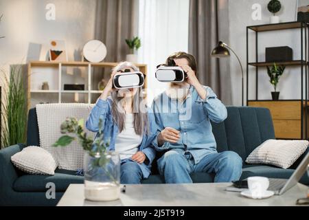 Angenehme alte Frau und ihr bärtiger Mann in VR-Brille sitzen auf dem Sofa im Wohnzimmer zu Hause. Lächelnde, reife Menschen verbringen Zeit mit elektronischen Geräten. Stockfoto