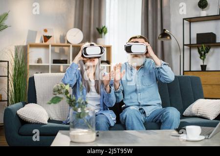 Glückliche, reife Familie, die zu Hause Spiele spielt und zu Hause VR-Brillen verwendet. Fröhliches Seniorenpaar, Familie sitzt auf der Couch, Sofa und unterhalten sich mit Innovationen. Stockfoto