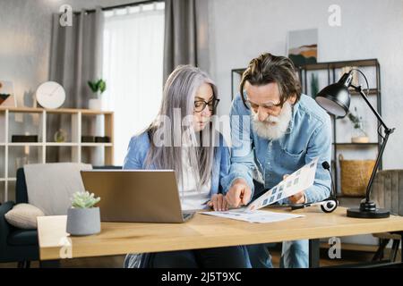 Reife Frau und bärtiger Mann Geschäftspartner in Casual Wear diskutieren gemeinsame Projekt in hellen Büro. Fröhliche ältere Kollegen, die moderne Gadgets für die Arbeit verwenden. Stockfoto