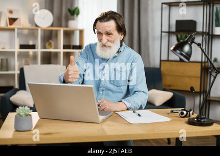 Der gutaussehende kaukasische Senior-Softwareentwickler in Freizeitkleidung tippt auf dem Laptop, während er am Arbeitsplatz sitzt und den Daumen nach oben zeigt. Beschäftigte Büroangestellte reifer Herr mit modernen Gadgets für die Arbeit Stockfoto
