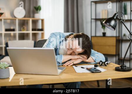 Erschöpfter, reifer kaukasischer Geschäftsmann schläft nach langem Papierkram auf dem Tisch. Müder älterer Mann mit grauem Bart in legerer Kleidung, der bei der Arbeit Pause einlegt. Überarbeiteter Begriff. Stockfoto