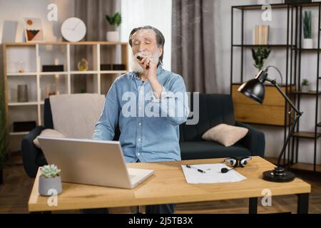 Elegant aussehender, gealterter, reifer Mann aus grauem Bart, Chef eines großen Unternehmens, der sich im Büro entspannt und Zigarre raucht, während er auf den Bildschirm eines Laptops blickt, während er am Tisch sitzt. Stockfoto