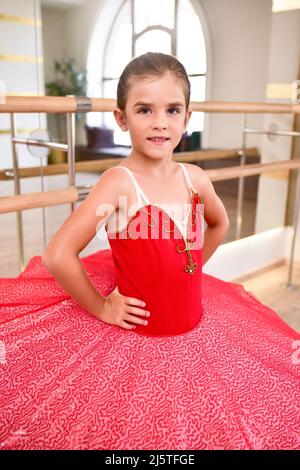 Niedliches Porträt einer schönen kleinen Ballerina in einem Performance-roten Kleid mit rosa Tutu. Sie lächelt, weil sie glücklich ist, ein Profi-Ball zu werden Stockfoto