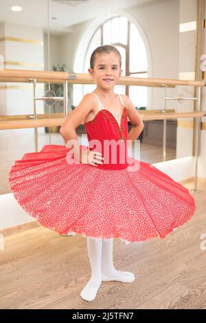Niedliches Porträt einer schönen kleinen Ballerina in einem Performance-roten Kleid mit rosa Tutu. Sie lächelt, weil sie glücklich ist, ein Profi-Ball zu werden Stockfoto