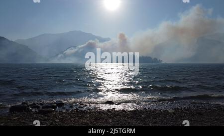 Sonnenlicht bricht durch den Rauch des Olgiasca-Waldbrands über dem Comer See, gesehen vom Ufer in Colico, Lecco, Lombardei, Italien, 18. April 2022 Stockfoto