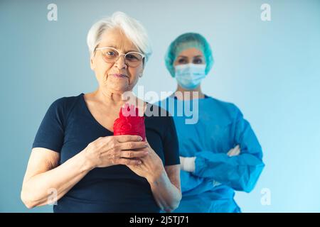 Ärztin, die hinter der dankbaren älteren europäischen Frau mit einem roten künstlichen Herzen, mittellange Aufnahme im Studio, blauer Hintergrund, Gesundheitskonzept steht. Hochwertige Fotos Stockfoto