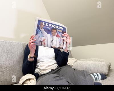 Paris, Frankreich - 25. Apr 2022: Frau liest auf einer gemütlichen Couch die neueste Zeitung Le Figaro deckt den amtierenden französischen Präsidenten Emmanuel Macron ab, nachdem sie Marine Le Pen für eine zweite fünfjährige Amtszeit geschlagen hatte Stockfoto