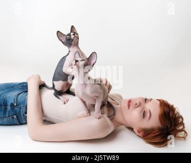 Zwei Vollblut-Kätzchen schauen nach oben, sitzen auf rothaariger junger Frau, die auf weißem Hintergrund auf ihrem Rücken liegt. Hübsche Frau mit kurzen Haaren trägt T-shir Stockfoto