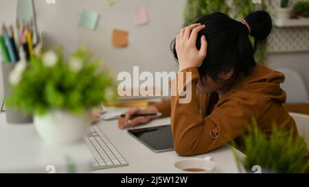 Gestresste und erschöpfte asiatische junge Grafikdesignerin, die an ihrem Büroarbeitsplatz arbeitet. Stockfoto