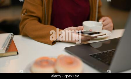 Nahaufnahme, Freiberuflerin Fernarbeit im Coworking Space des Cafés, mit Smartphone und Laptop-Computer, während sie Kaffee schlürft. Stockfoto