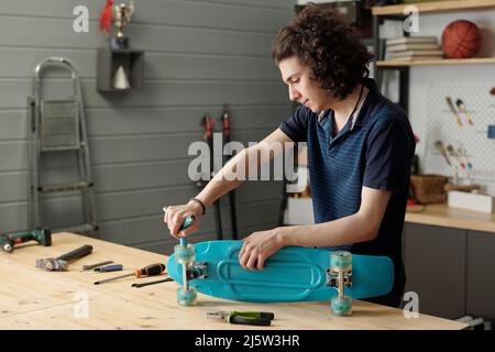 Jugendlicher Kerl mit Handwerkzeug, der Räder auf dem Skateboard montiert, während er am Holztisch in der Garage gegen die Stehleiter und andere Haushaltsgegenstände steht Stockfoto