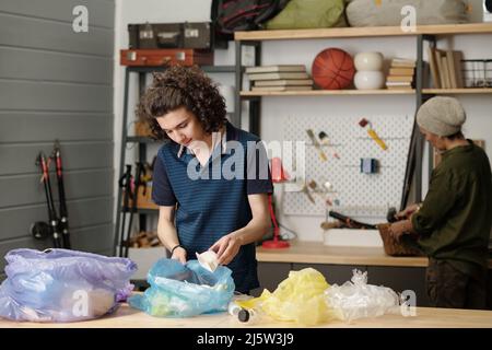 Jugendlicher Typ in der Casualwear, der Plastiklebensmittelbehälter in einen von mehreren Zellophansäcken legt, während er in der Garage Abfall nach Tisch sortiert Stockfoto