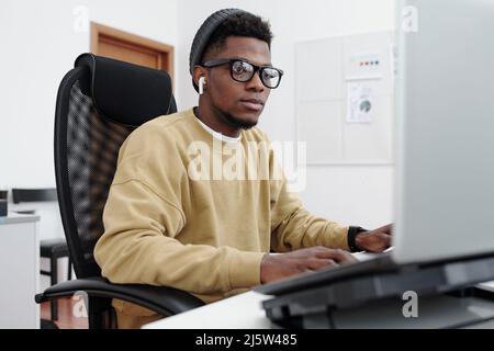 Junger moderner seriöser männlicher IT-Programmierer in gelbem Sweatshirt, der auf den Computerbildschirm schaut, während er im Büro Daten dekodiert Stockfoto