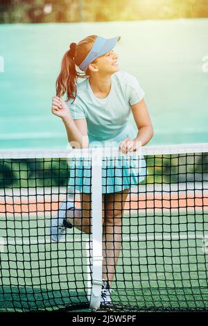 Sorgloses lächelndes Mädchen, das sich in hellblauer Sportkleidung auf einem Tennisplatznetz stützt. Zur Seite schauend, posiert. Beleuchtet mit abendlicher Sonneneinstrahlung. Stockfoto