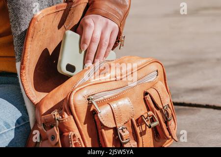 Crop anonyme Frau in Jacke und Jeans setzen Handy in stilvolle Leder-Handtasche, während auf sonnigen Straße in der Stadt sitzen Stockfoto