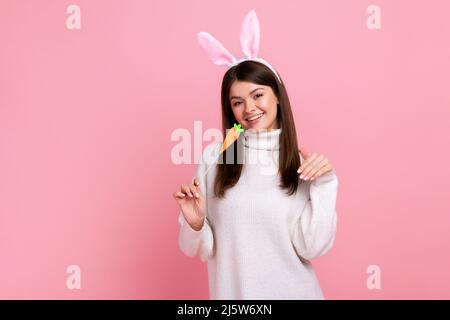 Lustige verspielte Mädchen mit Hasenohren stehen mit Karotte in den Händen und Blick auf die Kamera mit Lächeln, tragen weißen lässigen Stil Pullover. Innenaufnahme des Studios isoliert auf rosa Hintergrund. Stockfoto