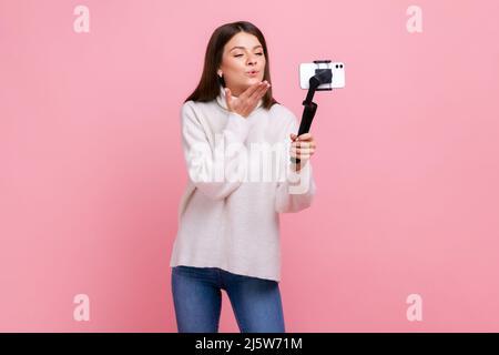 Happy Romantic hübsche Bloggerin, die Smartphone und Steadicam zum Streamen verwendet, sendet Luftküsse, trägt weißen Pullover im lässigen Stil. Innenaufnahme des Studios isoliert auf rosa Hintergrund. Stockfoto