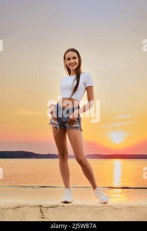 Fröhliche Frau in weißem T-Shirt und Jeans-Shorts, die auf einem Pier über dem herrlichen Sonnenuntergang posiert Stockfoto