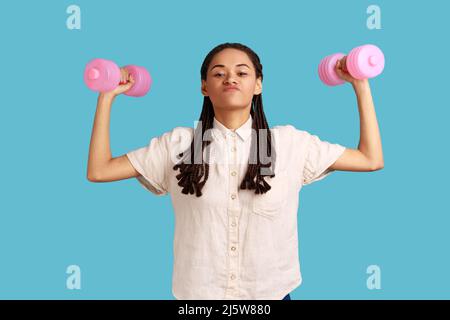 Selbstbewusste Frau mit schwarzen Dreadlocks, die die Muskeln mit Hanteln aufpumpt, selbstbewusst die Kamera anschaut und ein weißes Hemd trägt. Innenaufnahme des Studios isoliert auf blauem Hintergrund. Stockfoto