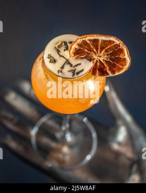 Orangefarbenes Cocktailglas mit einer Scheibe dehydrierter Orange, die auf einer handförmigen Metallskulptur ruht. Stockfoto