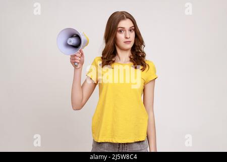 Verwirrt verwirrt verwirrt Teenager Mädchen mit braunen Haaren in gelben T-Shirt in den Händen halten Megaphon, kann keine gemeinsame Sprache zu finden, Blick auf die Kamera. Innenaufnahme des Studios isoliert auf grauem Hintergrund. Stockfoto